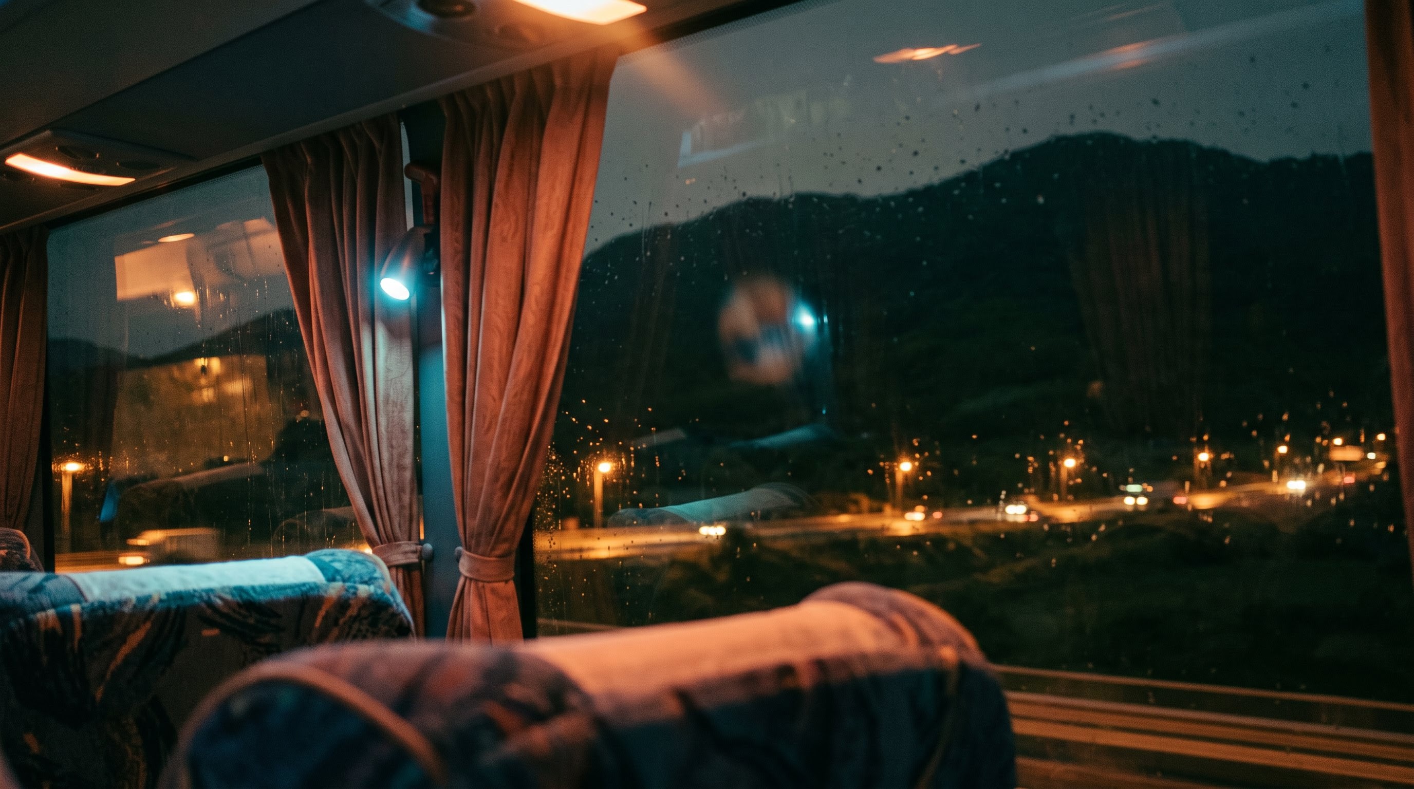 View from inside a sleeper bus at night, curtained window reflecting interior blue reading lights, dark green mountain silhouettes and scattered roadside lamps blurring past outside, rain-speckled glass, moody teal and amber grade, tight intimate framing over a seat back